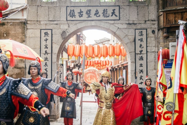 $vo['fname']|default='贵州安顺平坝区的天龙屯堡，是明代“调北征南”屯军后裔聚居地，被誉为“明代生活的活化石”。其历史可追溯至洪武十四年（1381年），朱元璋派军平定西南后，江浙汉族士兵在此屯田戍边，形成独特的军事移民文化。

图一：中式木构大门飞檐翘角，黑底金匾“天龙屯堡”端立，灯笼垂联、门楣面具，串起600年“调北征南”屯军记忆。
图二至四：金甲、黑红甲武士执枪列阵于“天龙屯堡古镇”石拱门前；红灯笼垂街，复现明代军傩遗风，活态演绎“寓兵于农”的屯堡精神。